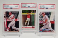 three 2018 Topps #300, 2019 Topps #100 & 2020 Topps #1 Mike Trout Trio Of 3 PSA 10'S baseball cards in a display case.